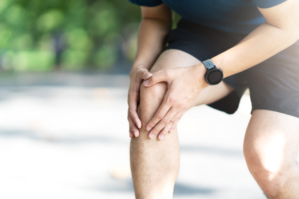 Young asian sporty man holding knee pain in running public nature park in the morning. Joint or leg injury in athlete or jogger concept with copy space. Millennial lifestyle runner with smart gadget.