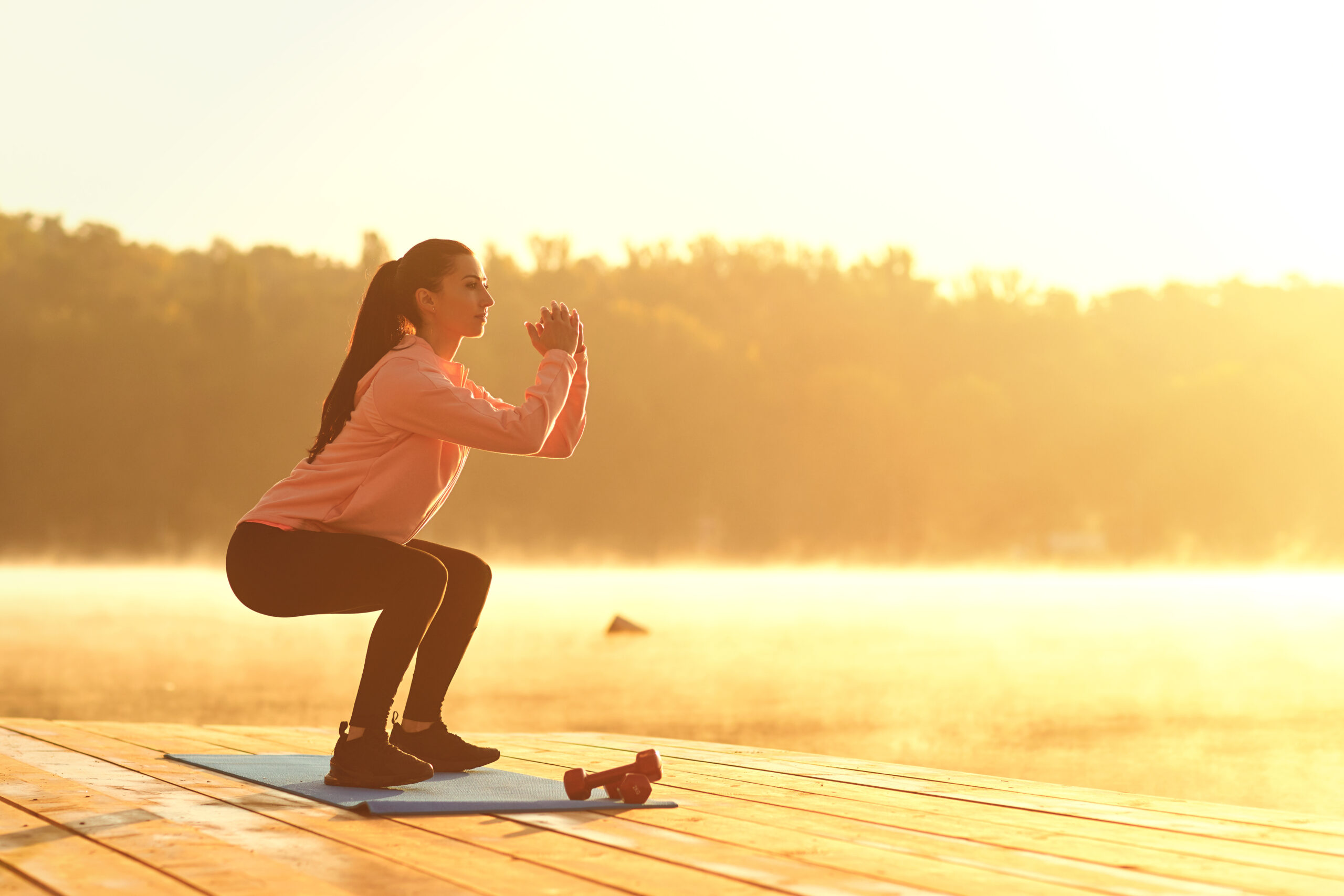 A young girl in training does squats at sunrise by the lake in the park in the summer in autumn.