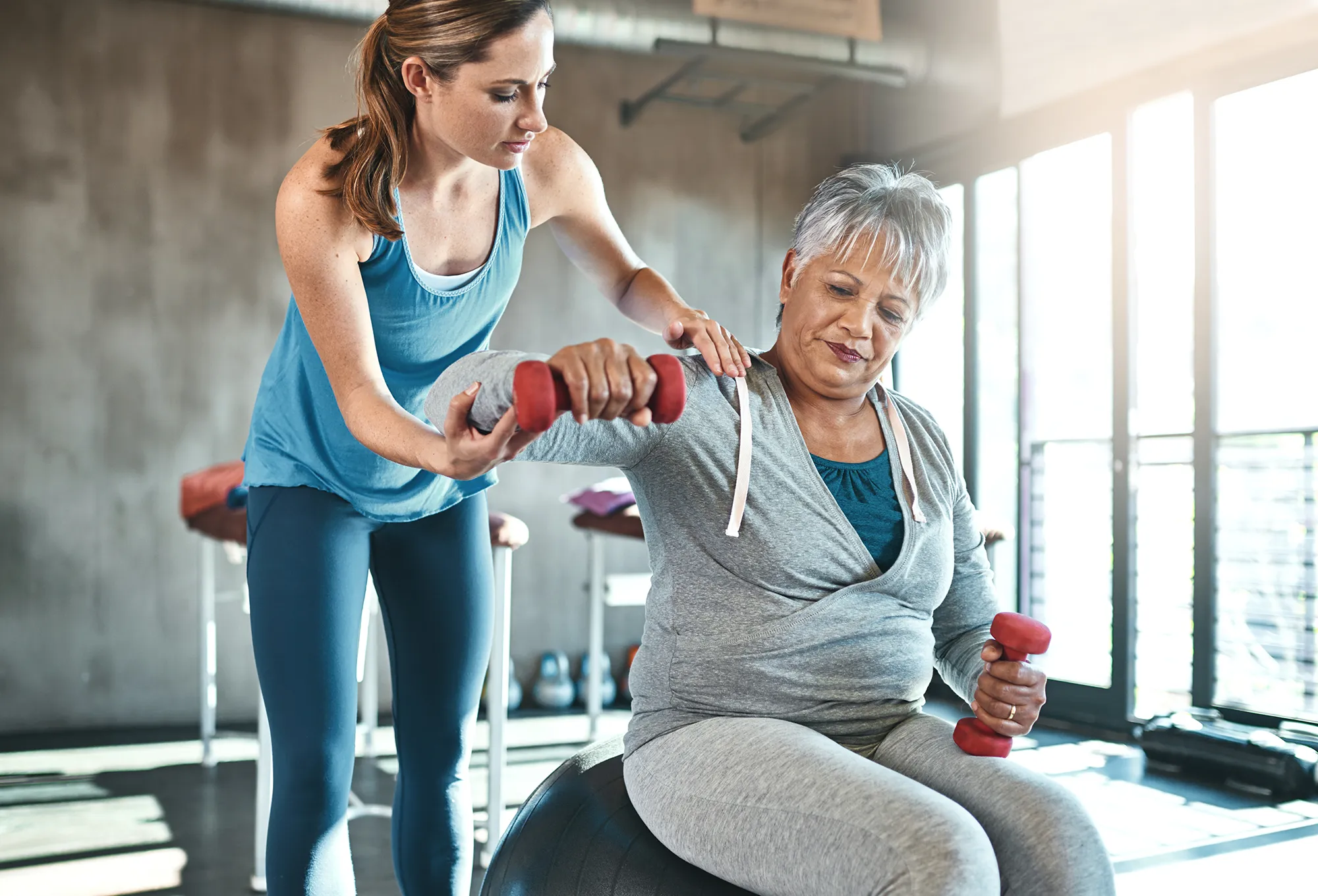 Senior woman, weights and helping with coach in gym for physio, arm exercise and rehabilitation on ball. Old female patient, instructor and workout equipment for arthritis with balance and dumbbells See Less