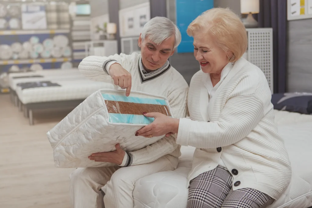 Cheerful senior man choosing orthopedic mattress, shopping with his wife at furniture store. lovely elderly couple examining orthopedic mattress sample, choosing new bed at home goods shop, copy space
