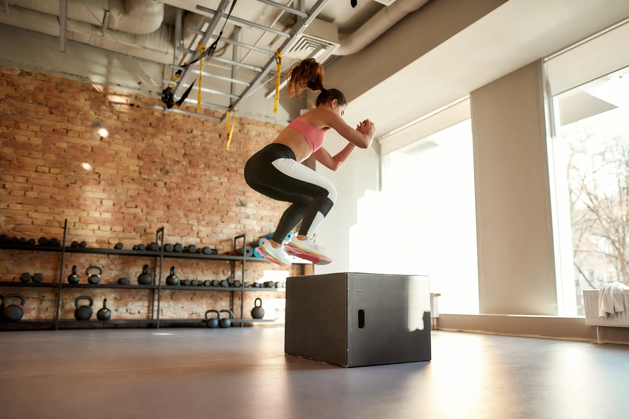 Sportive young woman jumping using plyo box while having workout at industrial gym
