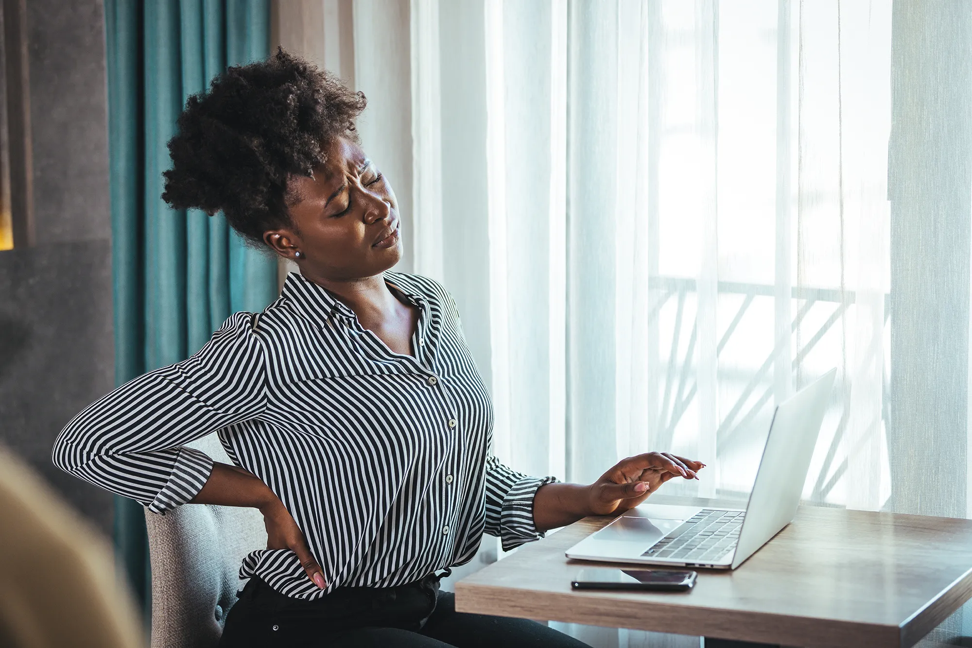 Tired african American millennial female worker sit at desk touch back suffer from lower spinal spasm, hurt unhealthy biracial woman stretch have strong backache, incorrect posture concept