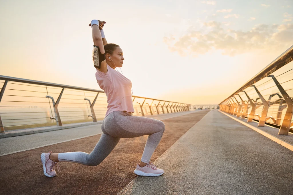 Determined young sportswoman exercising outdoor at dawn. Female athlete in sportswear stretching her arms up, performing squats and lunges, working out on city bridge in the early morning at sunrise.