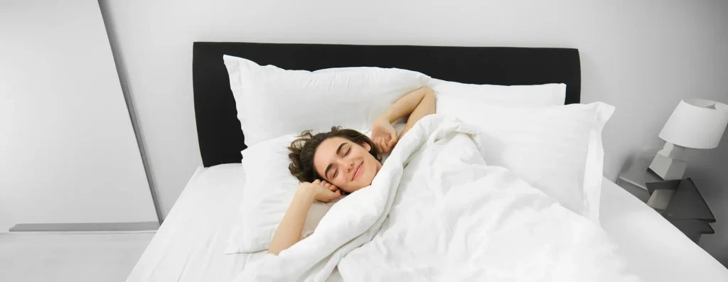 Portrait of smiling, pleased young woman stretching her arms, waking up after good night sleep, lying on soft white pillow in bed with comfortable linen sheets
