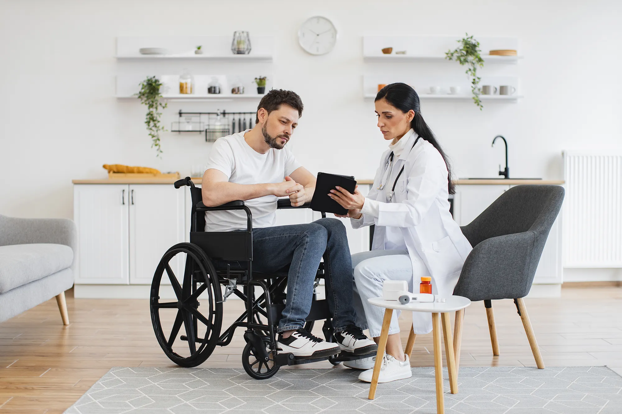 Full length shot of female brunette doctor checking up condition of bearded male patient at home. Nurse talking to mature man on wheelchair, showing examination results using modern tablet. 