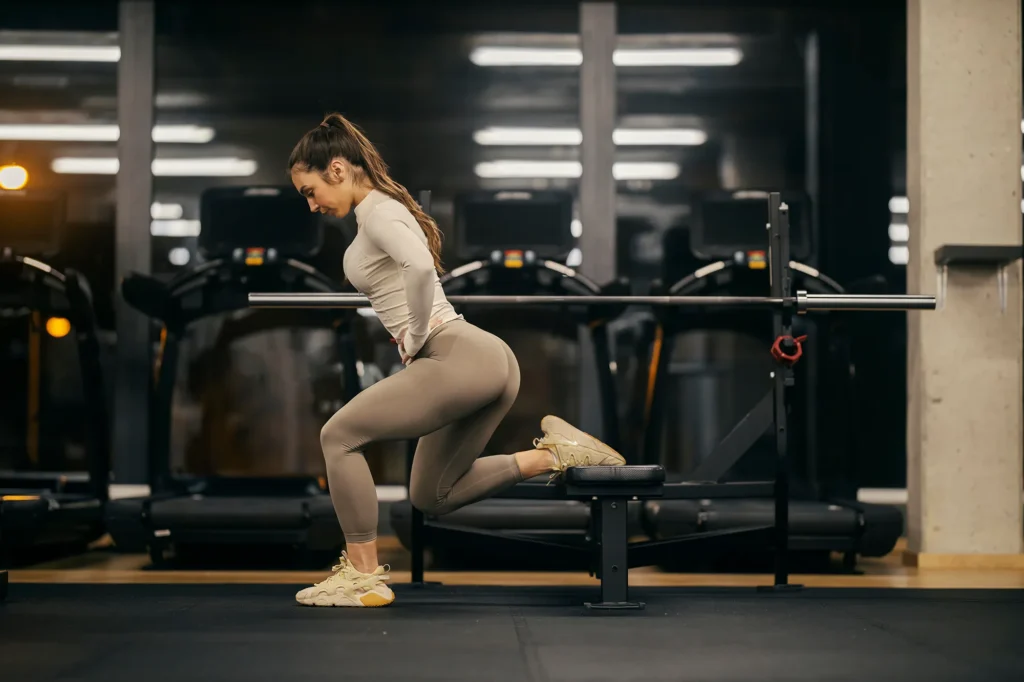 A sportswoman in shape is exercising split squats on a bench in a gym.
