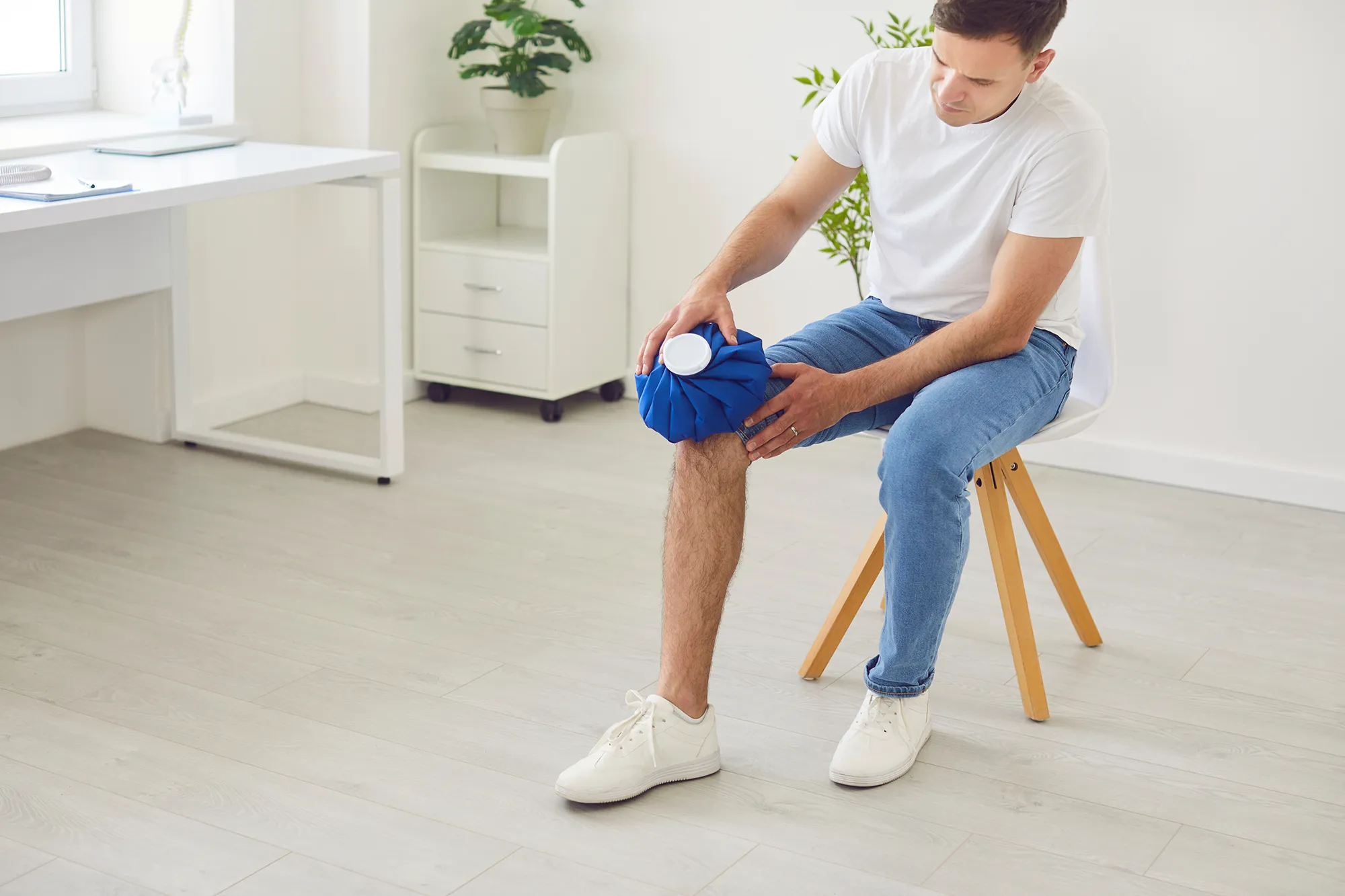  Portrait of a young man applying cold compress to leg sitting on chair at home. Male person holding ice pack bag on the knee to reduce pain. Inflammation, injury and health problems concept. 