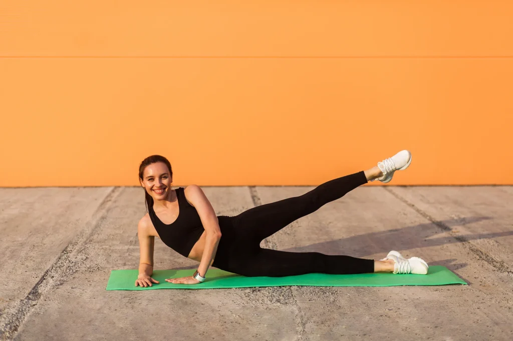 Cheerful athletic girl in tight sportswear, black pants and top, practicing yoga, doing side plank pose with leg lift, stretching muscles, training flexibility. Health care, sport activity outdoor 